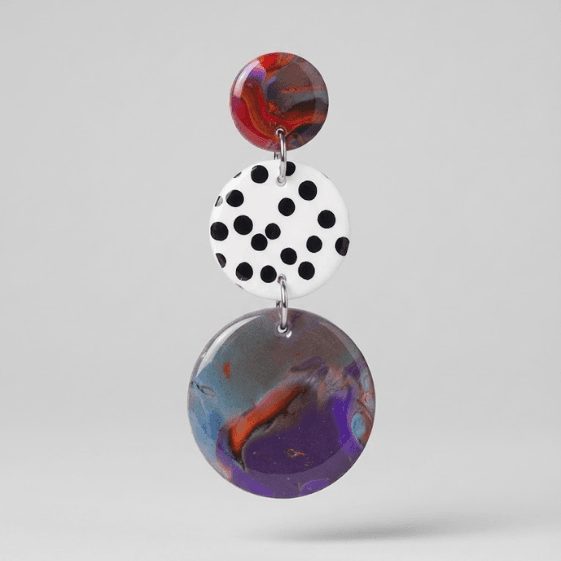 Close-up of marbled statement earrings with a red top disc, white disc with black polka dots, and a purple-blue marbled bottom disc on a light grey background.