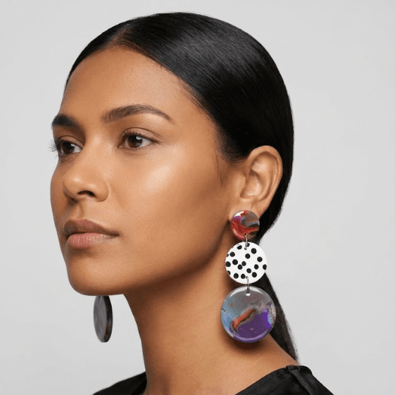 Model wearing marbled statement earrings with a red top disc, a white black polka dot middle disc, and a purple-blue marbled drop.