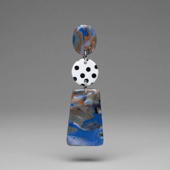 Close-up of handmade earrings with a marbled top disc, white disc with black polka dots, and a blue-grey marbled trapeze drop on a light grey background.