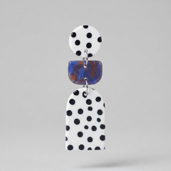 Close-up of polka dot statement earrings with a white round stud, blue marbled rectangle connector, and a white arch drop with black dots on a light grey background.