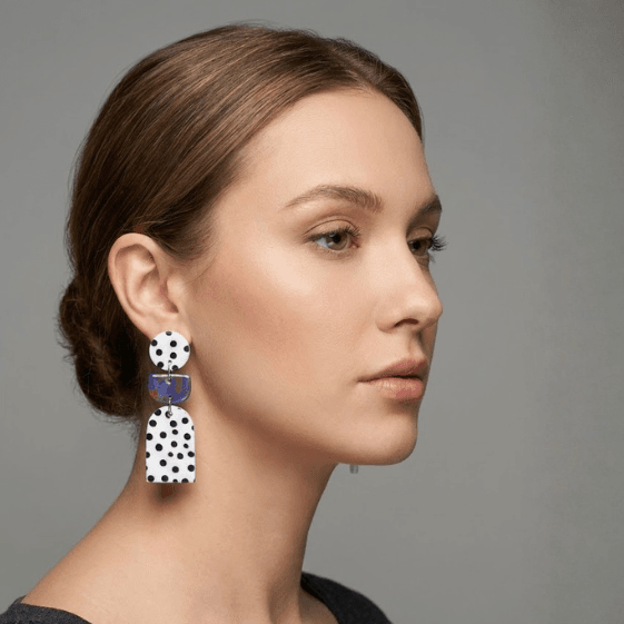 Model wearing polka dot statement earrings with a white round stud, blue marbled connector, and a white arch drop with black dots.