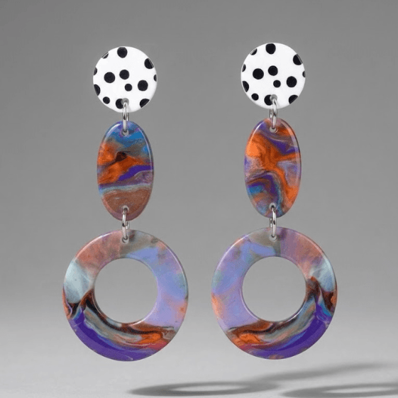 Close-up of bold earrings with a white black polka dot stud, orange-blue marbled oval connector, and large purple-pastel marbled hoop drop on a light grey background.