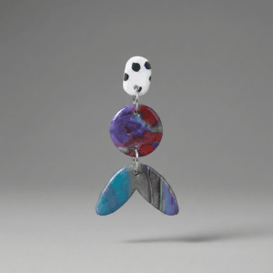 Close-up of statement earrings with a white dotted stud, purple-red marbled round disc, and teal-blue marbled tail-shaped drop on a light grey background.