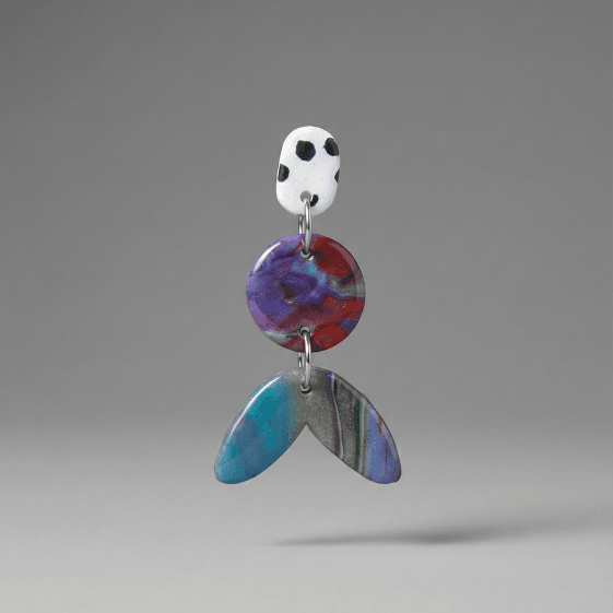 Close-up of statement earrings with a white dotted stud, purple-red marbled round disc, and teal-blue marbled tail-shaped drop on a light grey background.