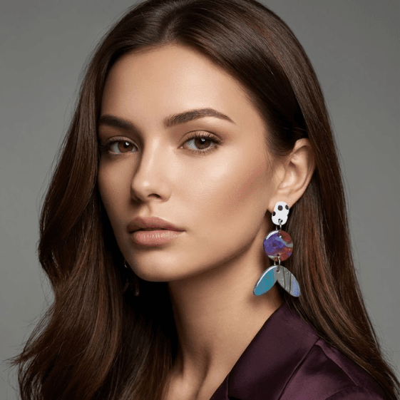 Model wearing statement earrings with a white dotted stud, a purple-red marbled round disc, and a teal-blue marbled tail-shaped drop.