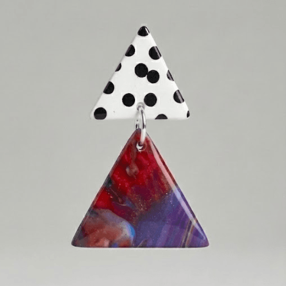 Close-up of dangle earrings with a white black polka dot triangle stud and a marbled purple-red triangle drop on a light grey background.