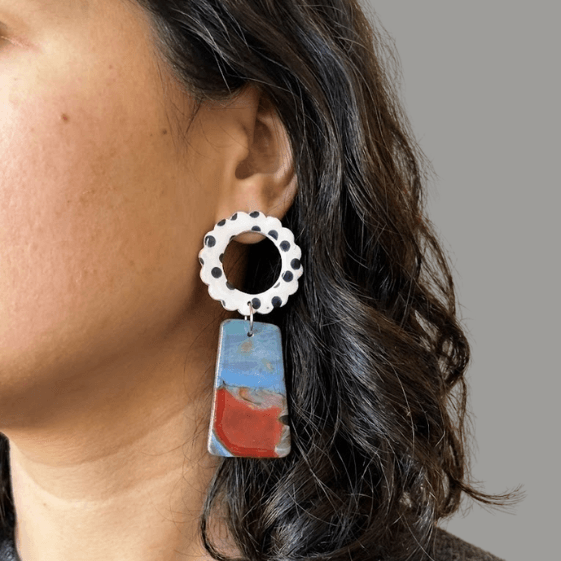 Model wearing flower earrings with a white scalloped ring top with black dots and a marbled blue-red trapeze drop.