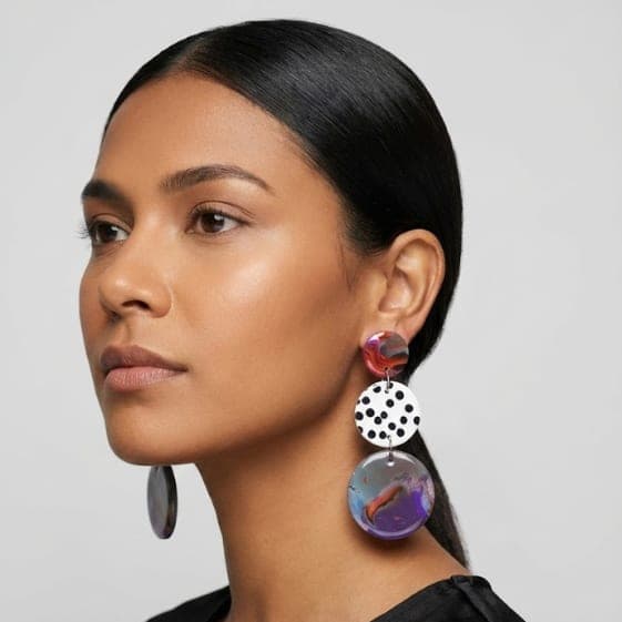 Model wearing marbled statement earrings with a red top disc, a white black polka dot middle disc, and a purple-blue marbled drop.