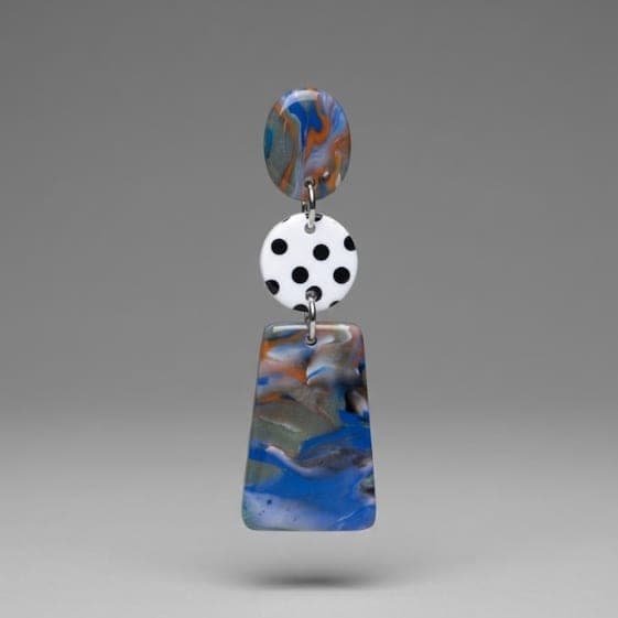 Close-up of handmade earrings with a marbled top disc, white disc with black polka dots, and a blue-grey marbled trapeze drop on a light grey background.