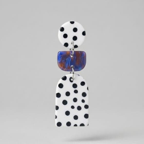 Close-up of polka dot statement earrings with a white round stud, blue marbled rectangle connector, and a white arch drop with black dots on a light grey background.