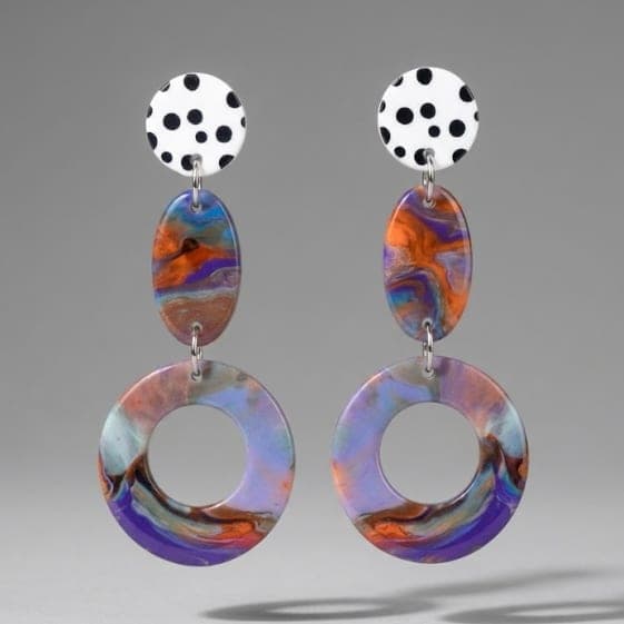 Nahaufnahme von Statement-Ohrringen mit weiß-schwarzem Polka-Dot-Stecker, orange-blau marmoriertem ovalen Verbinder und großem lila-pastell marmorierten Hoop-Anhänger vor hellgrauem Hintergrund.