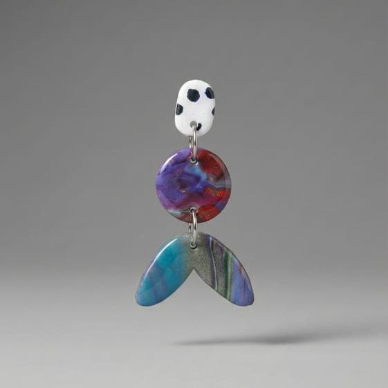 Close-up of statement earrings with a white dotted stud, purple-red marbled round disc, and teal-blue marbled tail-shaped drop on a light grey background.
