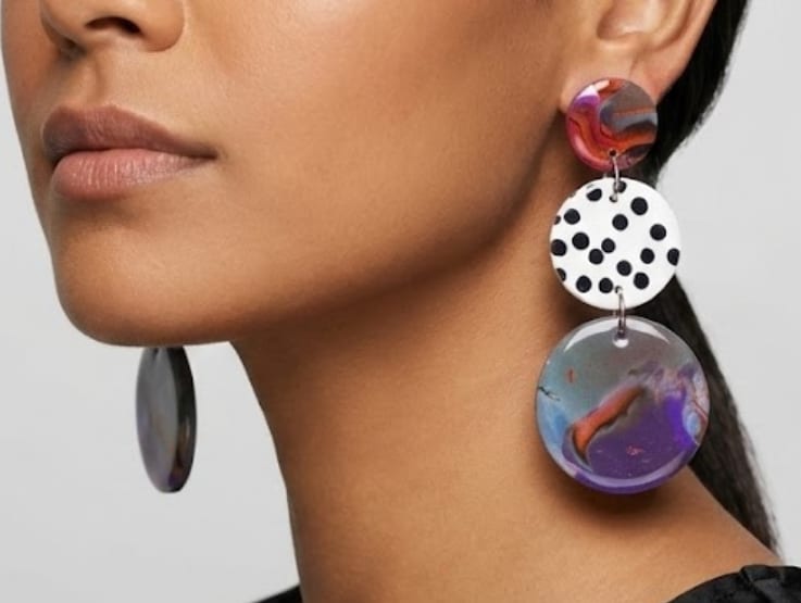 Woman wearing abstract sculptural earrings featuring a black and white polka dot stud and a large marbled colorful flower drop.